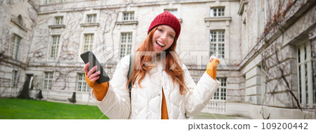 Cheerful redhead girl winning, celebrating victory, read great news on mobile phone and jumping from joy, saying yes, standing outside building in yard 109200442