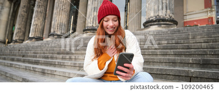 Young smiling woman with long red hair, sits near building on stairs with smartphone, using mobile application, sending message Young smiling woman with long red hair, sits near building on stairs with smartphone, using mobile application, sending message 109200465