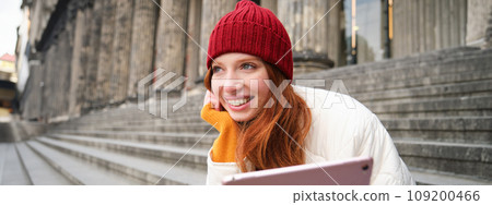 Outdoor shot of young stylish redhead girl sits on staircase and connects to public wifi, uses digital tablet, reads news on gadget Outdoor shot of young stylish redhead girl sits on staircase and connects to public wifi, uses digital tablet, reads news on gadget 109200466