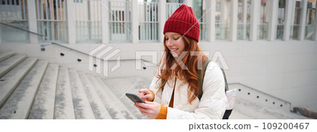 Smiling redhead girl, student tourist walks around city, goes up the stairs, looks at mobile phone map to get around town, sends message on smartphone Smiling redhead girl, student tourist walks around city, goes up the stairs, looks at mobile phone map to get around town, sends message on smartphone 109200467