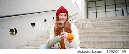 Smiling traveler, redhead girl tourist sits on stairs with flask, drinks hot coffee from thermos while travelling and sightseeing around foreign city, sits on stairs and rests Smiling traveler, redhead girl tourist sits on stairs with flask, drinks hot coffee from thermos while travelling and sightseeing around foreign city, sits on stairs and rests 109200469