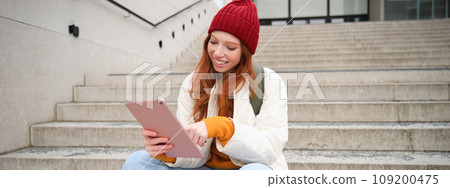 Happy stylish redhead girl, student in red hat, holds digital tablet, uses social media app, searches something online, connects to wifi Happy stylish redhead girl, student in red hat, holds digital tablet, uses social media app, searches something online, connects to wifi 109200475