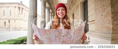 Adventurous redhead girl walks in town with paper map, explores city as tourist, looks for popular tourism attractions, looks around excited and smiles Adventurous redhead girl walks in town with paper map, explores city as tourist, looks for popular tourism attractions, looks around excited and smiles 109200683