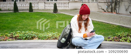 Young stylish female student with backpack, sits on bench and uses smartphone, plays on mobile phone 109200690