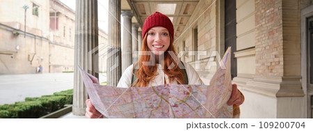 Smiling young redhead woman in red hat, looks at paper map to look for tourist attraction. Tourism and people concept. Girl explores city, tried to find way 109200704