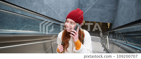 Portrait of young redhead woman walks around city, goes up stairs with mobile phone, talks on smartphone and smiles 109200707