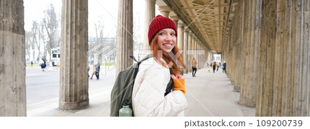 Female tourist with red long hair, holds backpack with thermos, turns back at camera with happy smile, explores city and popular tourism landmarks Female tourist with red long hair, holds backpack with thermos, turns back at camera with happy smile, explores city and popular tourism landmarks 109200739