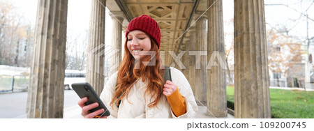 Mobile broadband and people. Smiling redhead 20s girl with backpack, uses smartphone on street, holds mobile phone and looks at application Mobile broadband and people. Smiling redhead 20s girl with backpack, uses smartphone on street, holds mobile phone and looks at application 109200745