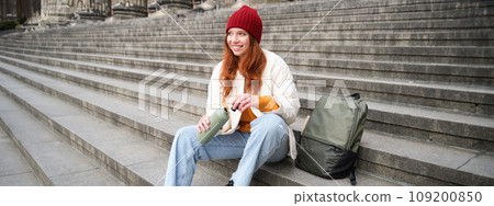 Smiling tourist, girl sits on stairs, rests on staircase, takes thermos from backpack, drinks hot coffee from flask 109200850