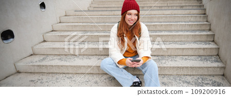Hipster ginger girl, redhead woman sits on stairs with smartphone, waits for someone and messages on social media on mobile phone app Hipster ginger girl, redhead woman sits on stairs with smartphone, waits for someone and messages on social media on mobile phone app 109200916