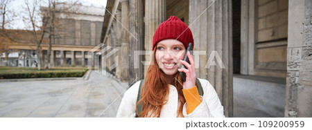 Smiling redhead female tourist talks on mobile phone and walks around city. Happy student in red hat calls friend, stands on street and uses smartphone 109200959