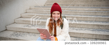 Young stylish girl, redhead female students sits on stairs outdoors with digital tablet, reads, uses social media app on gadget, plays games while waits on street 109200981