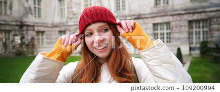 Portrait of smiling redhead woman puts on red hat and smiles, wearing warm clothes while exploring city streets Portrait of smiling redhead woman puts on red hat and smiles, wearing warm clothes while exploring city streets 109200994