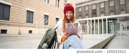 Redhead girl smiles, sits outdoors near building with digital tablet, thermos and backpack, connects to public internet and searches smth online on her gadget Redhead girl smiles, sits outdoors near building with digital tablet, thermos and backpack, connects to public internet and searches smth online on her gadget 109201022