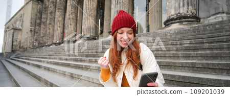 Achievement celebration. Happy redhead girl sits on stairs outdoors and looks at phone, triumphs, wins something and looks satisfied at mobile screen 109201039