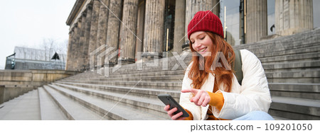 Stylish young redhead woman, talking on mobile phone app, using social media application, looking for something online on smartphone, sits on stairs outdoors Stylish young redhead woman, talking on mobile phone app, using social media application, looking for something online on smartphone, sits on stairs outdoors 109201050