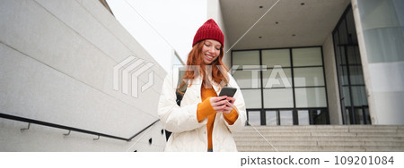 Redhead girl, young woman tourist with backpack, holds smartphone, looks for route on mobile application, searches for hotel on phone map, smiles happily Redhead girl, young woman tourist with backpack, holds smartphone, looks for route on mobile application, searches for hotel on phone map, smiles happily 109201084
