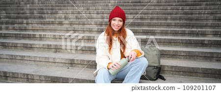 Smiling tourist, girl sits on stairs, rests on staircase, takes thermos from backpack, drinks hot coffee from flask Smiling tourist, girl sits on stairs, rests on staircase, takes thermos from backpack, drinks hot coffee from flask 109201107