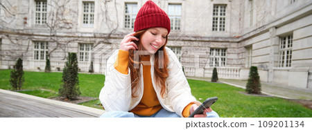 Young smiling redhead girl sits on bench and uses smartphone app, reads news online, watches video on mobile phone while relaxed in park 109201134