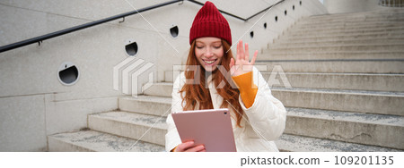 Joyful redhead teen girl, student with digital tablet, says hello, waves hand at gadget camera, connects to video chat, talks to friend in application Joyful redhead teen girl, student with digital tablet, says hello, waves hand at gadget camera, connects to video chat, talks to friend in application 109201135