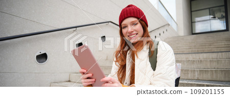 Happy stylish redhead girl, student in red hat, holds digital tablet, uses social media app, searches something online, connects to wifi Happy stylish redhead girl, student in red hat, holds digital tablet, uses social media app, searches something online, connects to wifi 109201155