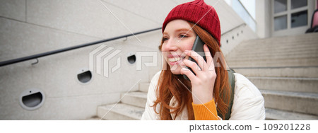 Young stylish redhead girl in red hat, sits on street and talks on mobile phone, has telephone conversation, rings her friend while relaxes outdoors Young stylish redhead girl in red hat, sits on street and talks on mobile phone, has telephone conversation, rings her friend while relaxes outdoors 109201228