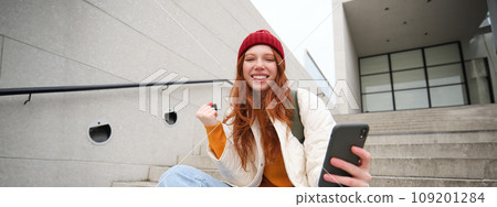 Joyful young redhead girl sits on street stairs, say yes, wins and celebrates with phone in hand, receive positive news on her mobile application and rejoice 109201284