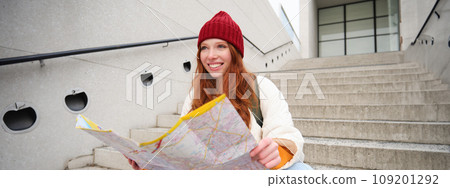 Young smiling redhead girl, tourist sits on stairs outdoors with city paper map, looking for direction, traveller backpacker explores city and looks for sightseeing 109201292