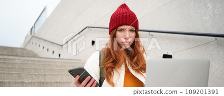 Portrait of redhead woman sits on stairs, uses laptop and holds smartphone, looks confused and upset at computer screen, tries to connect to wifi public internet 109201391