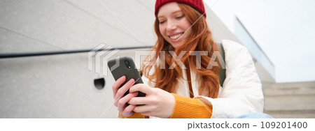 Mobile phones and people. Young stylish redhead girl sits on stairs with telephone, uses smartphone app, reads smth online 109201400