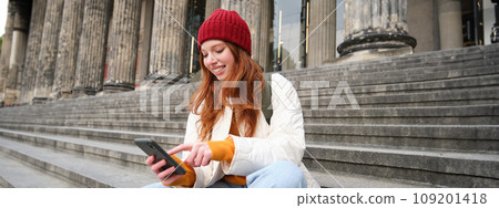 Portrait of young urban girl in red hat, sits on stairs near museum, holds mobile phone, connects to public wifi and surfs net, uses smartphone apps Portrait of young urban girl in red hat, sits on stairs near museum, holds mobile phone, connects to public wifi and surfs net, uses smartphone apps 109201418
