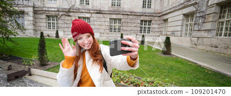 Portrait of girl blogger records video and says hi at phone camera. Young woman takes selfie with smartphone app, waves hand, video chats in application Portrait of girl blogger records video and says hi at phone camera. Young woman takes selfie with smartphone app, waves hand, video chats in application 109201470
