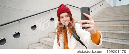 Mobile phone and people lifestyle. Stylish redhead girl takes selfie on her smartphone, poses for photo with mobile phone in hand, smiles happily, sits on stairs outdoors Mobile phone and people lifestyle. Stylish redhead girl takes selfie on her smartphone, poses for photo with mobile phone in hand, smiles happily, sits on stairs outdoors 109201478