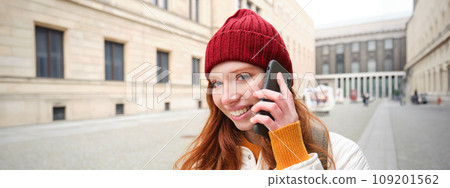 Mobile broadband and people. Smiling young redhead woman walks in town and talks on mobile phone, calling friend on smartphone, using internet to make a call abroad 109201562