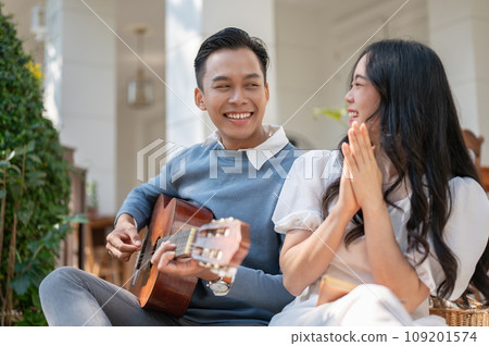 Cheerful and happy young Asian couple enjoying playing guitar and picnic at home backyard together. Cheerful and happy young Asian couple enjoying playing guitar and picnic at home backyard together. 109201574