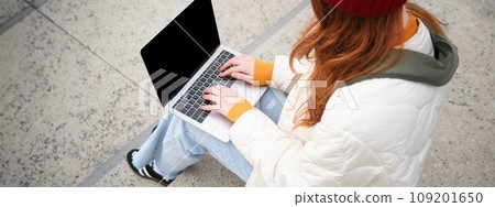 Rear view of girl student typing on laptop keyboard, blank black screen for website application advertisement, sitting on stairs outdoors, connects to public wifi Rear view of girl student typing on laptop keyboard, blank black screen for website application advertisement, sitting on stairs outdoors, connects to public wifi 109201650