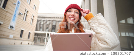 Happy redhead girl in red hat, walks around city with digital tablet, connects to public internet wifi and looks for route, looks at map on her gadget Happy redhead girl in red hat, walks around city with digital tablet, connects to public internet wifi and looks for route, looks at map on her gadget 109201707