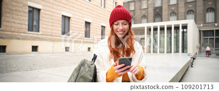 Young redhead woman with smartphone, sitting outdoors with backpack, student looking at her mobile phone, texts message Young redhead woman with smartphone, sitting outdoors with backpack, student looking at her mobile phone, texts message 109201713