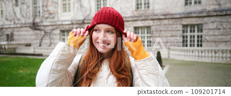 Portrait of smiling redhead woman puts on red hat and smiles, wearing warm clothes while exploring city streets Portrait of smiling redhead woman puts on red hat and smiles, wearing warm clothes while exploring city streets 109201714