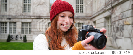 Redhead girl photographer takes photos on professional camera outdoors, captures streetstyle shots, looks excited while taking pictures 109201749