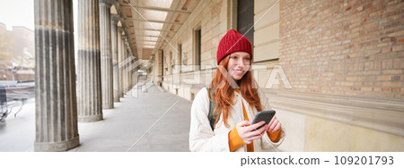 Smiling redhead girl in red hat, looks at map on smartphone app, checks her location, searches for a hostel. Tourist searches for popular attractions on mobile phone Smiling redhead girl in red hat, looks at map on smartphone app, checks her location, searches for a hostel. Tourist searches for popular attractions on mobile phone 109201793