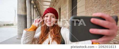 Cute young redhead woman takes selfie on street with mobile phone, makes a photo of herself with smartphone app on street 109201813