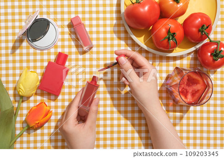 Advertising scene for tomato-based cosmetics. Lipstick, perfume bottle, mirrors and fresh tomatoes are decorated on a checkered background. The woman's hand is holding a tube of tomato red lipstick 109203345