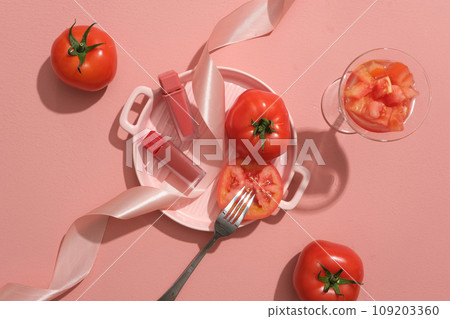 Top view of red ripe tomatoes displayed on pink background. A pink ceramic pan container lipsticks decorated with pink ribbon and fork. Creative scene for advertising. Mockup for design 109203360