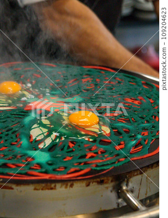 Asian multicolored pancakes made of layers of red and green. The cook breaks a raw egg into dough. Close-up Asian multicolored pancakes made of layers of red and green. The cook breaks a raw egg into dough. Close-up 109204623