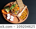 Wooden menagerie with healthy snacks. Pieces of fresh vegetables, roasted chickpeas and grain loaves. Close-up on a black background 109204625