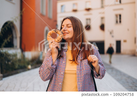 Attractive young female 30s tourist with backpack is holding prezel, traditional polish snack on the Market square in Krakow. Traveling Europe in spring. Selective focus. High quality photo Attractive young female 30s tourist with backpack is holding prezel, traditional polish snack on the Market square in Krakow. Traveling Europe in spring. Selective focus. High quality photo 109204758
