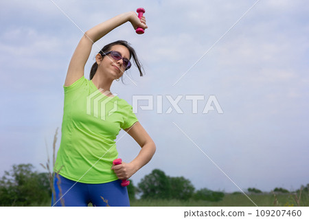 Beautiful hispanic woman in bright colored sportswear doing exercise with pink dumbbells in her hands in the city park on background blue sky. Outdoors healthy lifestyle concept, sports, fitness, yoga 109207460