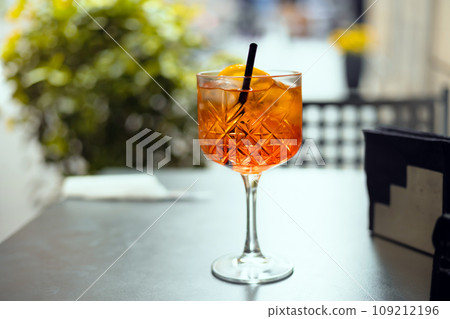 Aperol Spritz Cocktail. Alcoholic beverage based on table with ice cubes and oranges outdoors. Served cocktail with orange slice and straw placed on the table of sidewalk cafe in Italy 109212196