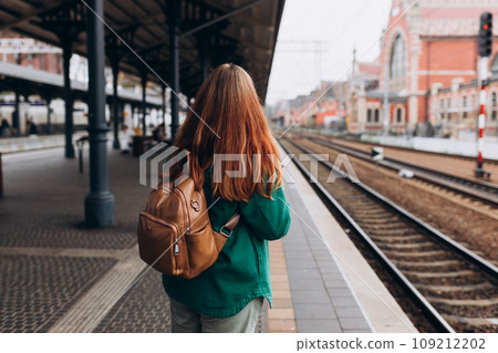 Back view of a young woman traveler with small backpack on the railway station, 30s women waiting for train. Tourism, solo vacation, rear view, no face 109212202